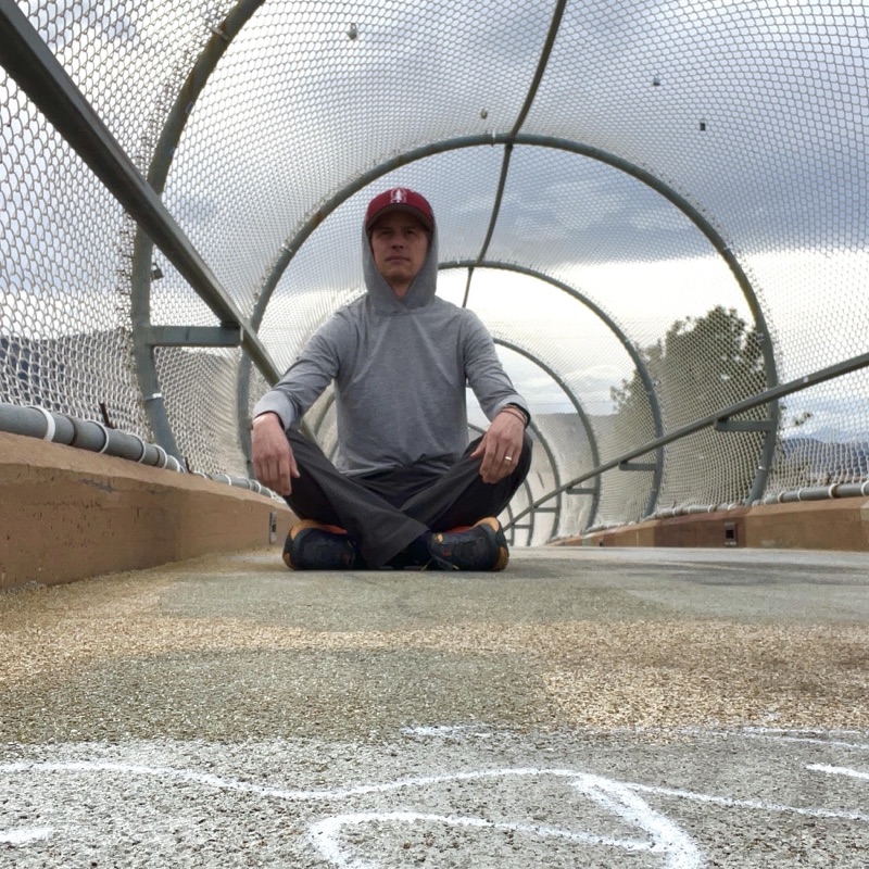 Nate sitting cross-legged in a mesh-covered footbridge, wearing a gray hoodie and a red cap